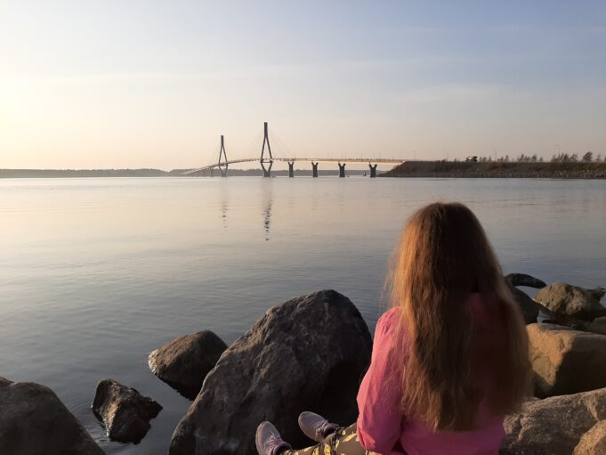 Flicka sitter på sten vid vattenbrynet. Bro i bakgrunden. Tyttö istuu merne rannalla. taustalla silta.